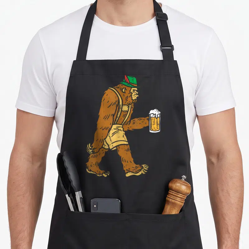 Bigfoot german apron - Orange orangutan walking in yellow overalls and green hat holding beer mug — shown on a black cotton canvas apron against a white background