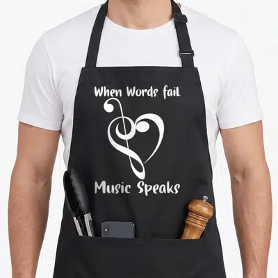 Musical music quotes apron - White treble clef shaped like a heart with text When Words fail Music Speaks — shown on a black cotton canvas apron against a white background