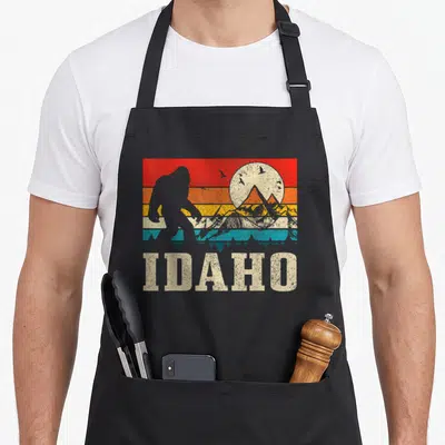 Hunter camping apron - Retro Idaho Bigfoot silhouette against red orange and teal sunset stripes with IDAHO text — shown on a black cotton canvas apron against a white background