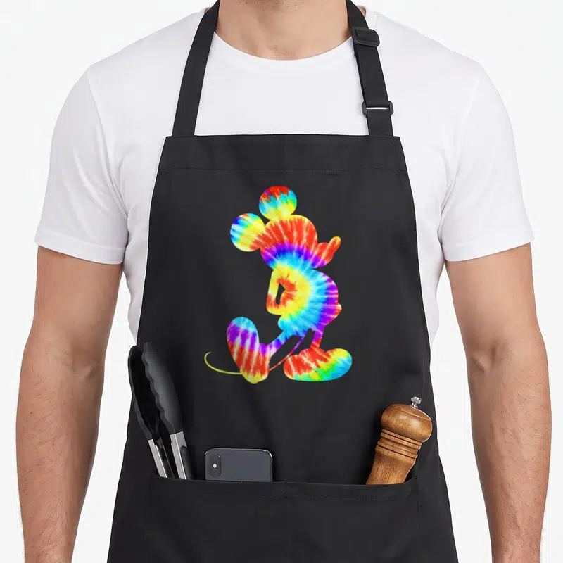 Dye tie apron - Mickey Mouse silhouette filled with red yellow green blue purple tie-dye pattern — shown on a black cotton canvas apron against a white background