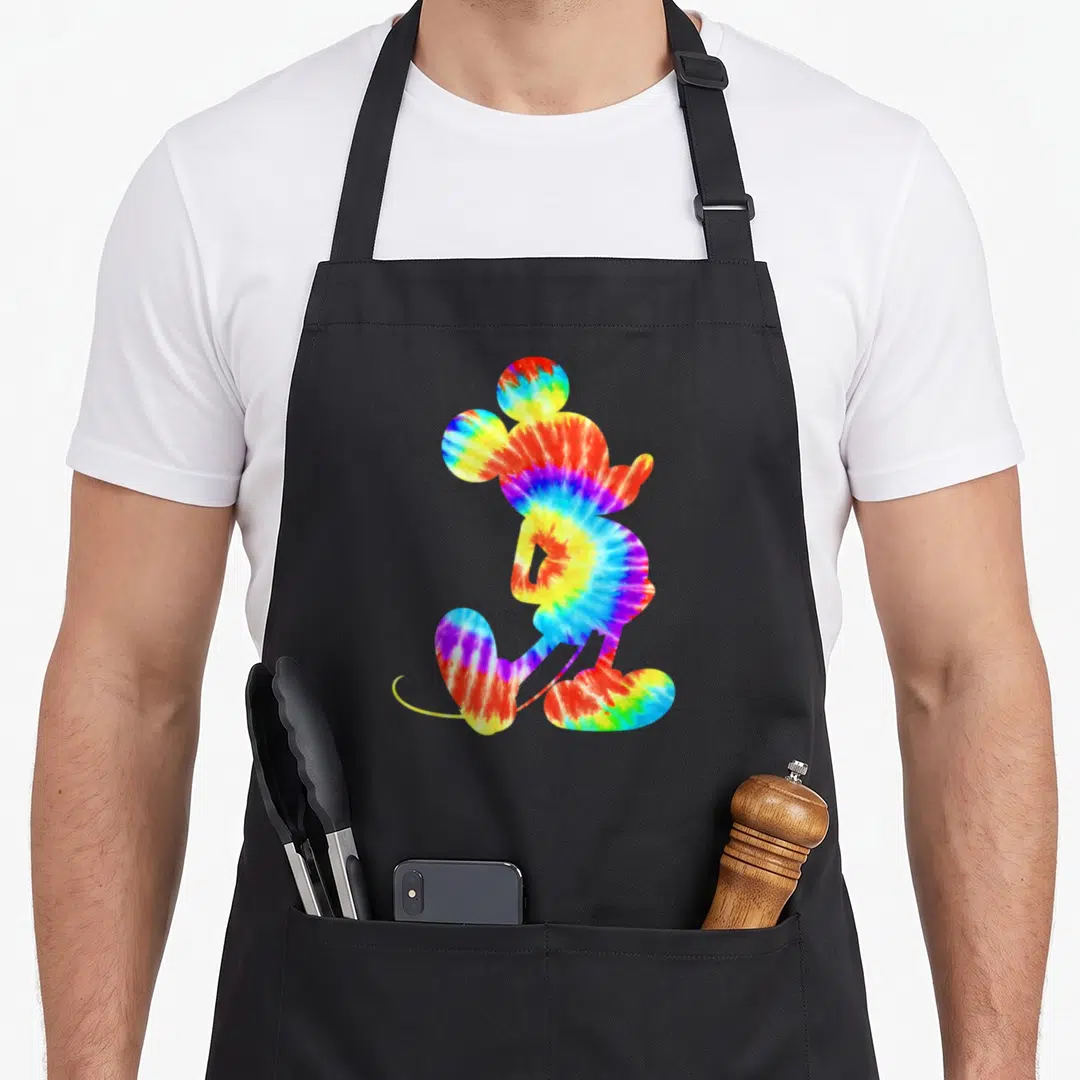 Dye tie apron - Mickey Mouse silhouette filled with red yellow green blue purple tie-dye pattern — shown on a black cotton canvas apron against a white background