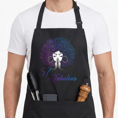 57th hair apron - Woman with word-art afro in purple and blue, hands together, text 57 & Fabulous below — shown on a black cotton canvas apron against a white background