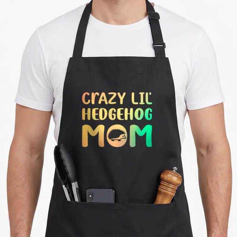 Cute lil crazy hedgehog lady apron - CRAZY LIL HEDGEHOG MOM text with hedgehog silhouette in orange yellow green — shown on a black cotton canvas apron against a white background