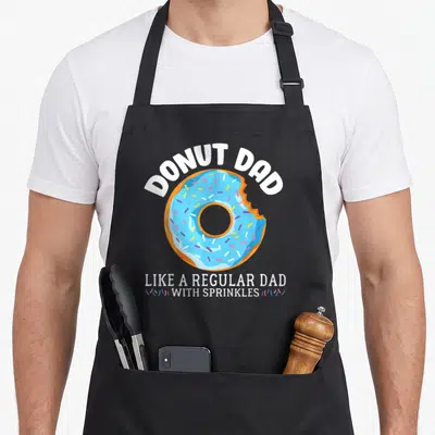 Father sprinkle apron - Blue frosted donut with bite mark and white cartoon text about sprinkles and regular dad — shown on a black cotton canvas apron against a white background