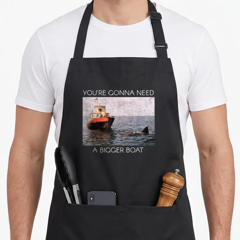 Screen boat apron - You're Gonna Need A Bigger Boat text above and below boat and shark fin photo — shown on a black cotton canvas apron against a white background