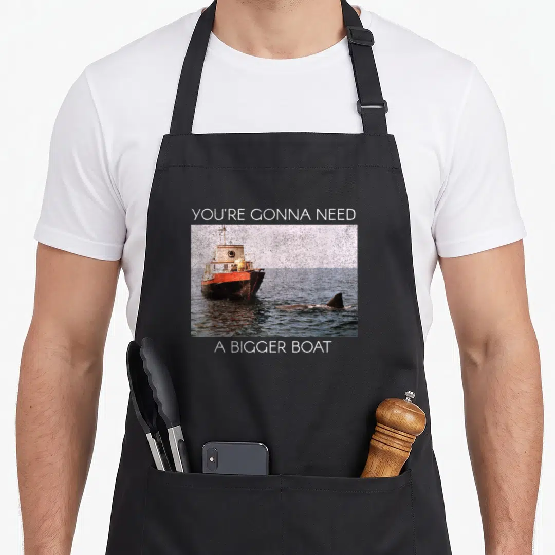 Screen boat apron - You're Gonna Need A Bigger Boat text above and below boat and shark fin photo — shown on a black cotton canvas apron against a white background
