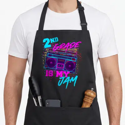 Jam 2nd apron - Retro boombox with 2ND GRADE IS MY JAM text and colorful splatters on yellow triangle — shown on a black cotton canvas apron against a white background