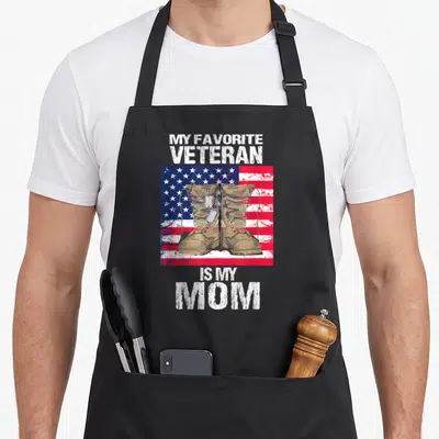 Veteran proud mother's day aprons - Tan combat boots with dog tags on a distressed US flag background with white text — shown on a black cotton canvas apron against a white background