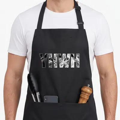 Yahweh lions apron - YHWH text filled with a close-up lion face illustration in black and white — shown on a black cotton canvas apron against a white background