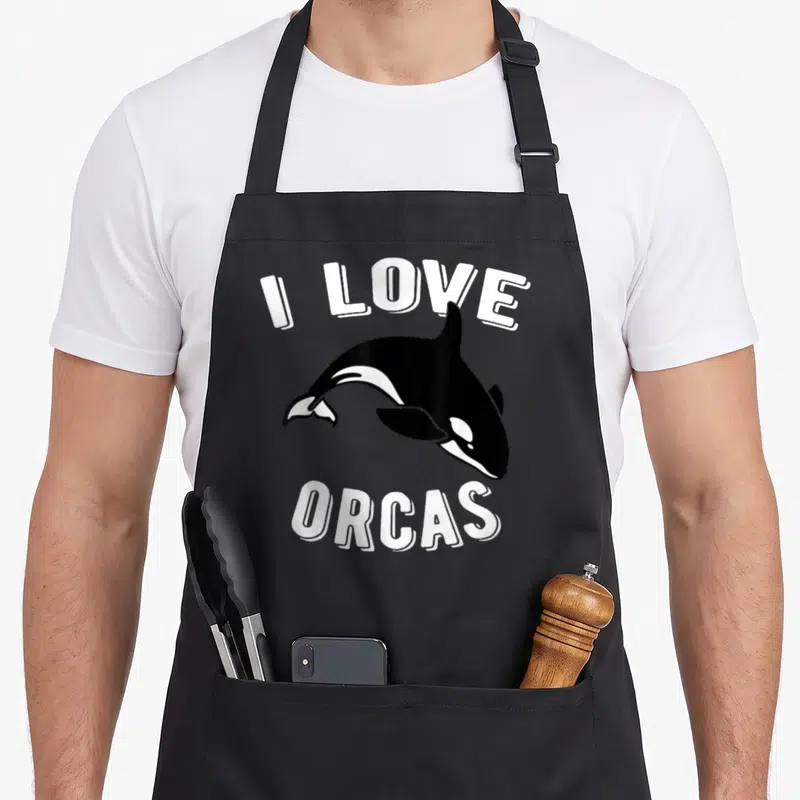 Orcas killer whale apron - I LOVE ORCAS text arching over and under a cartoon orca in black and white — shown on a black cotton canvas apron against a white background
