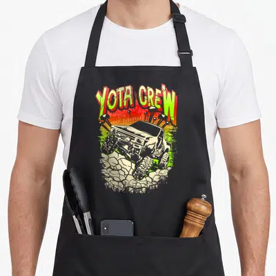 Funny rock crawler girl apron - YOTA CREW text in lime green and red above a truck on rocks with gradient background — shown on a black cotton canvas apron against a white background