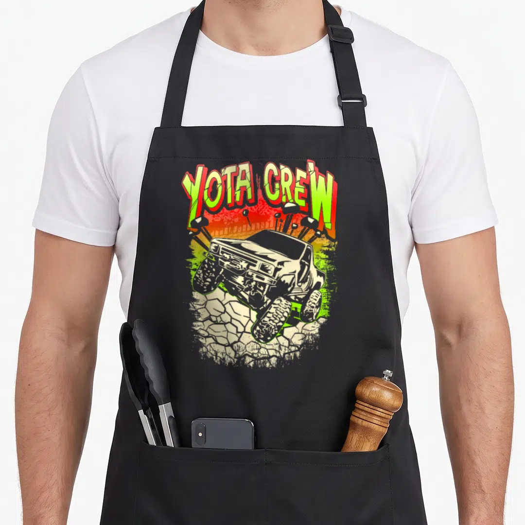 Funny rock crawler girl apron - YOTA CREW text in lime green and red above a truck on rocks with gradient background — shown on a black cotton canvas apron against a white background