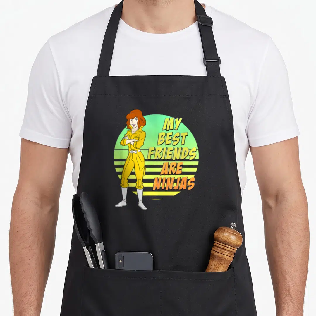 Oneil april apron - Cartoon woman in yellow jumpsuit beside My Best Friends Are Ninjas text on green striped circle — shown on a black cotton canvas apron against a white background