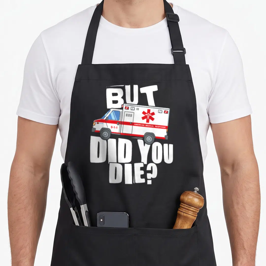 But paramedic apron - Cartoon ambulance with white red blue text reading BUT AMERICAN MEDICAL RESPONSE 9-1-1 DID YOU DIE — shown on a black cotton canvas apron against a white background