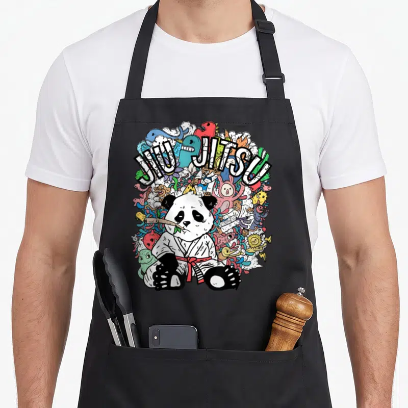 Panda jitsu apron - Cartoon panda in gi with JIU JITSU text surrounded by colorful monsters — shown on a black cotton canvas apron against a white background