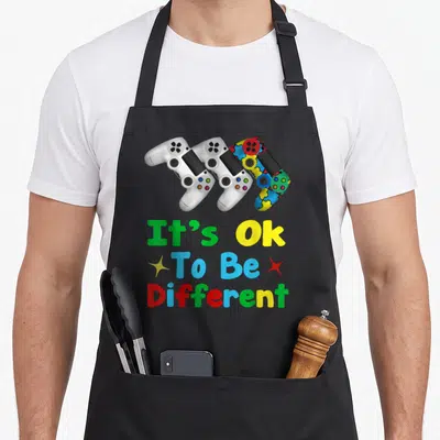 Autism game aprons - Three game controllers including one with puzzle pieces above It's Ok To Be Different text — shown on a black cotton canvas apron against a white background