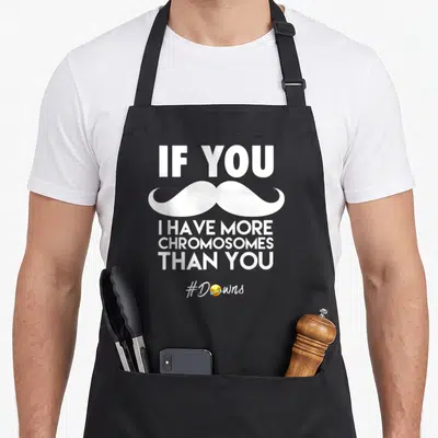 Mustache chromosomes apron - White text IF YOU I HAVE MORE CHROMOSOMES THAN YOU with mustache and laughing emoji hashtag — shown on a black cotton canvas apron against a white background