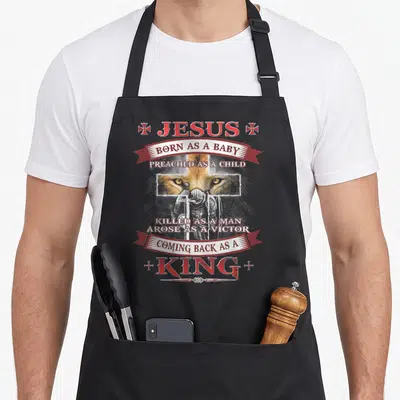 Baby christian apron - Lion face and knight with sword surrounded by JESUS BORN AS A BABY text in red and gold — shown on a black cotton canvas apron against a white background