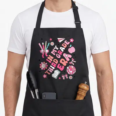 Third groovy grade apron - IN MY THIRD GRADE ERA text with cartoon school supplies, flowers, bows, and a checkered — shown on a black cotton canvas apron against a white background