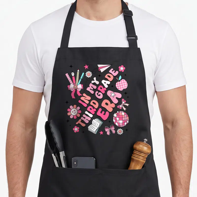 Third groovy grade apron - IN MY THIRD GRADE ERA text with cartoon school supplies, flowers, bows, and a checkered — shown on a black cotton canvas apron against a white background