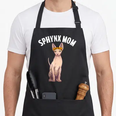 Sphynx grandma apron - Sitting pink sphynx cat with yellow pizza bow and white SPHYNX MOM text — shown on a black cotton canvas apron against a white background