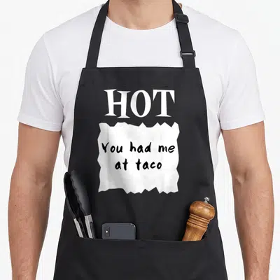 Hot sauce halloween apron - Torn white paper graphic with You had me at taco text below large HOT letters in black — shown on a black cotton canvas apron against a white background