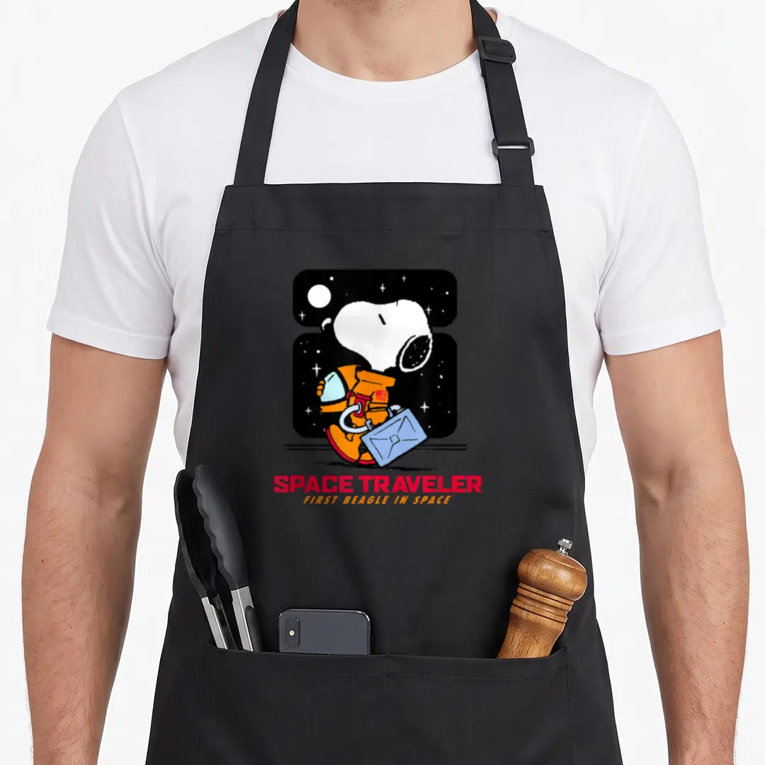 Space traveler apron - Snoopy in orange spacesuit holding blue briefcase with red and orange text — shown on a black cotton canvas apron against a white background