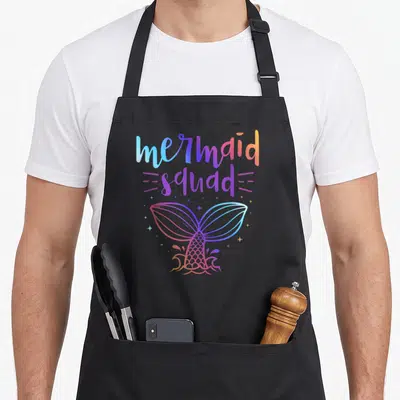 Vintage mermaid birthday apron - Gradient mermaid squad text above a colorful tail with scattered stars — shown on a black cotton canvas apron against a white background