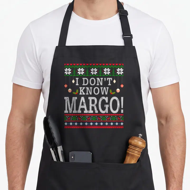 Margo ugly christmas sweater apron - I DON'T KNOW MARGO text with snowflakes reindeer and holly in white red and green — shown on a black cotton canvas apron against a white background