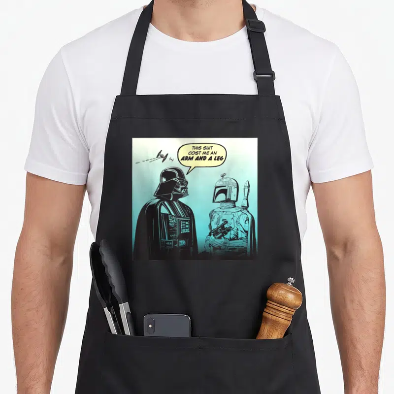Comic boba apron - Darth Vader and Boba Fett line art with speech bubble text on teal background — shown on a black cotton canvas apron against a white background