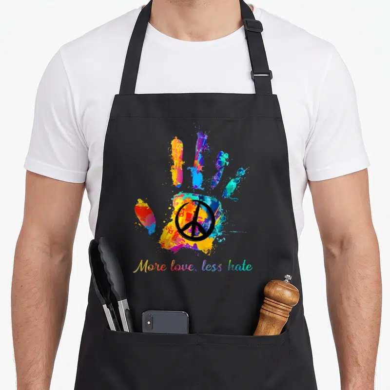 Hippie handprint apron - Multicolored watercolor hand with black peace sign and rainbow text — shown on a black cotton canvas apron against a white background