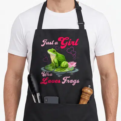 Cool frog aprons - Green frog on lily pad with pink lotus and hearts. Text Just a Girl Who Loves Frogs in pink script — shown on a black cotton canvas apron against a white background