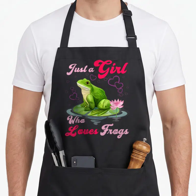 Cool frog aprons - Green frog on lily pad with pink lotus and hearts. Text Just a Girl Who Loves Frogs in pink script — shown on a black cotton canvas apron against a white background