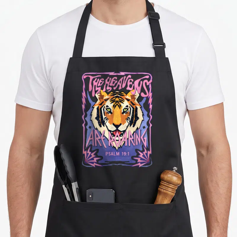 Heaven tiger retro apron - Cartoon tiger head with pink text THE HEAVENS ARE ROARING PSALM 19:1 and lightning bolts — shown on a black cotton canvas apron against a white background