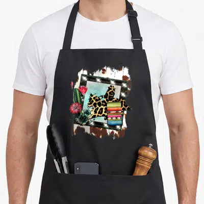 Barrel cheetah print apron - Leopard silhouette in light-up frame with cactus, succulent, and striped blanket — shown on a black cotton canvas apron against a white background
