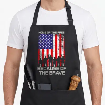 Free patriotic apron - Distressed USA flag with soldier silhouettes and text Home of the Free Because of the Brave — shown on a black cotton canvas apron against a white background