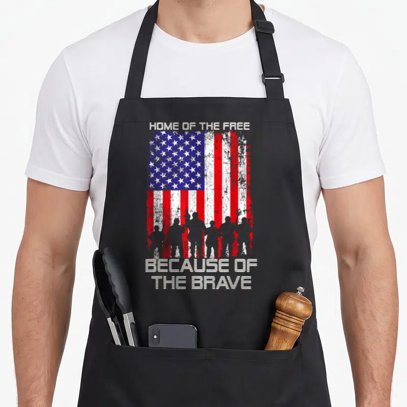 Free patriotic apron - Distressed USA flag with soldier silhouettes and text Home of the Free Because of the Brave — shown on a black cotton canvas apron against a white background
