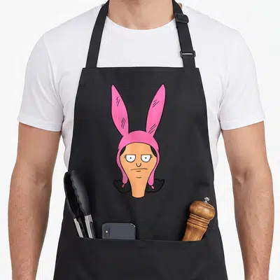 Louise bobs burger apron - Cartoon character head wearing pink bunny hat with peach skin and black hair — shown on a black cotton canvas apron against a white background