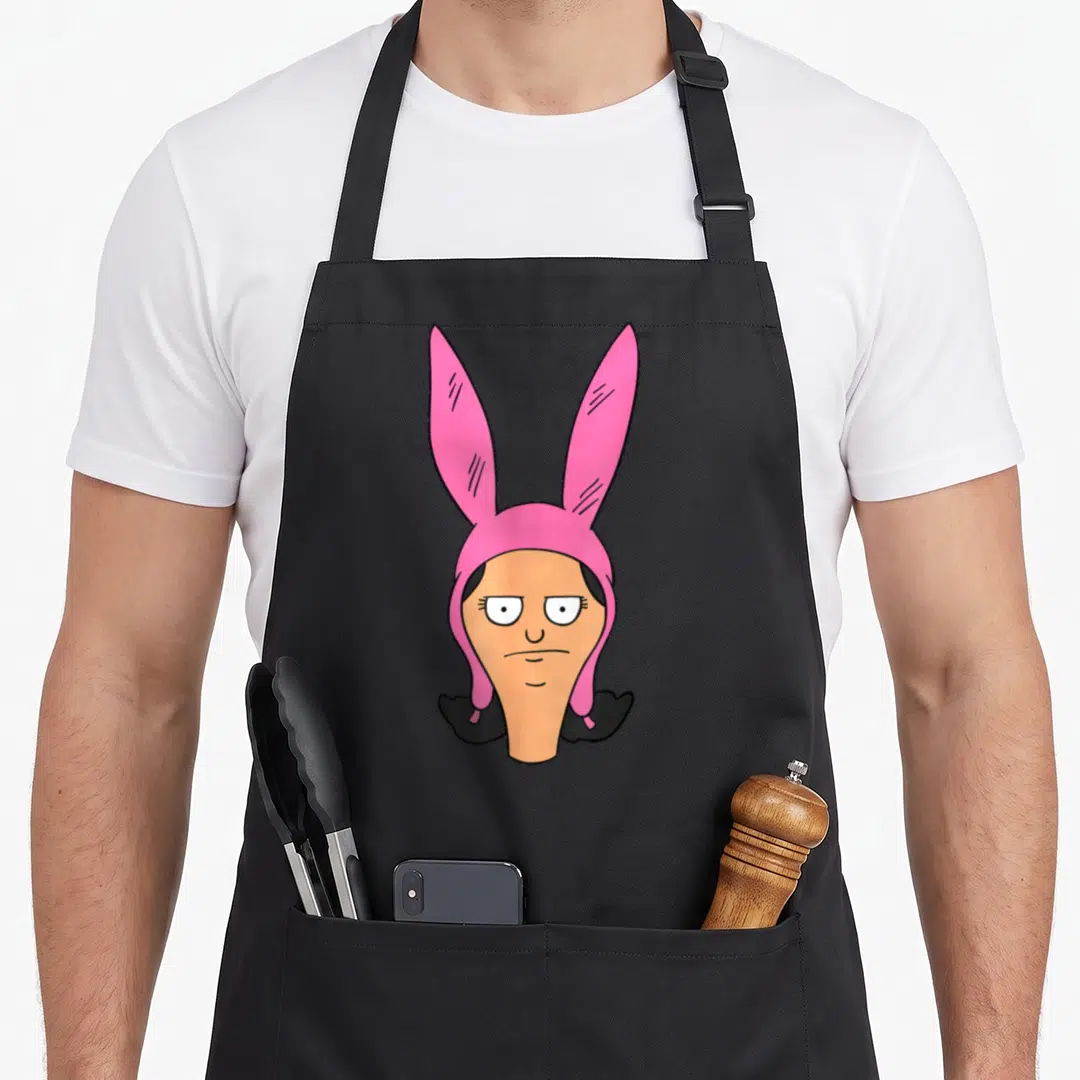 Louise bobs burger apron - Cartoon character head wearing pink bunny hat with peach skin and black hair — shown on a black cotton canvas apron against a white background