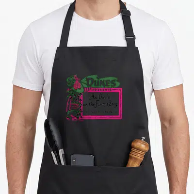Dune hotel aprons - Retro Dunes Las Vegas sign with palm trees and An Oasis script in green pink black — shown on a black cotton canvas apron against a white background