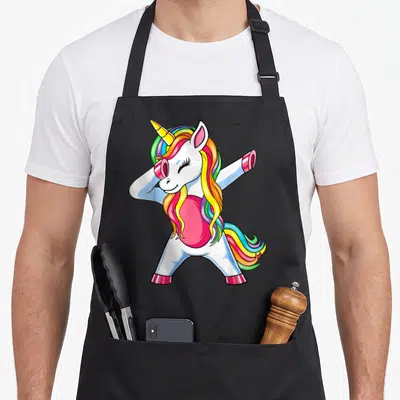 Funny dabbing rainbow apron - Cartoon unicorn dabbing with rainbow mane in white pink yellow blue green — shown on a black cotton canvas apron against a white background
