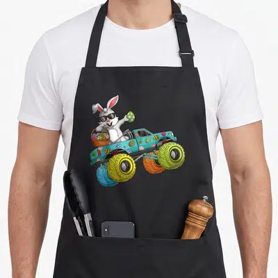 Dabbing easter apron - Cartoon rabbit in teal monster truck holding eggs and basket with four large colorful wheels — shown on a black cotton canvas apron against a white background