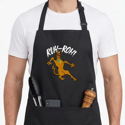 Ruh roh decade apron - Brown cartoon dog with white RUH-ROH! text and cyan collar — shown on a black cotton canvas apron against a white background