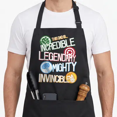 Legendary father day apron - Stacked text THIS DAD IS INCREDIBLE LEGENDARY MIGHTY INVINCIBLE with colored superhero — shown on a black cotton canvas apron against a white background