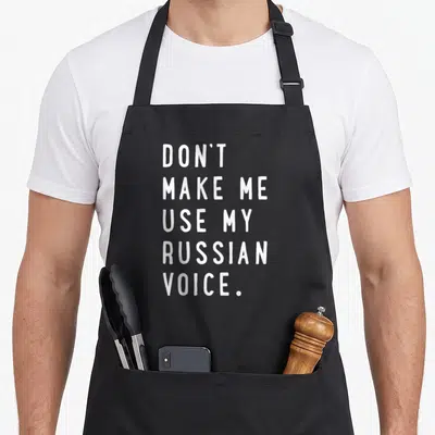 Use russian apron - White text reading DON'T MAKE ME USE MY RUSSIAN VOICE stacked vertically — shown on a black cotton canvas apron against a white background