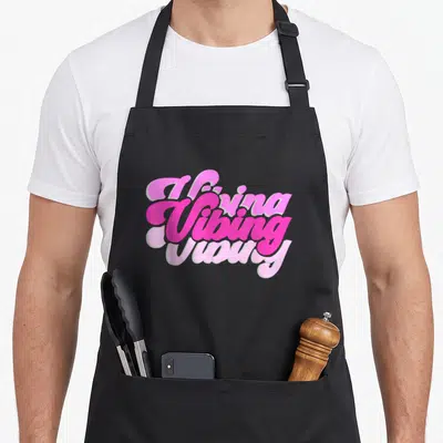 Funny vibing novelty apron - Stacked pink cursive text reading Vibing in three shades on black fabric — shown on a black cotton canvas apron against a white background