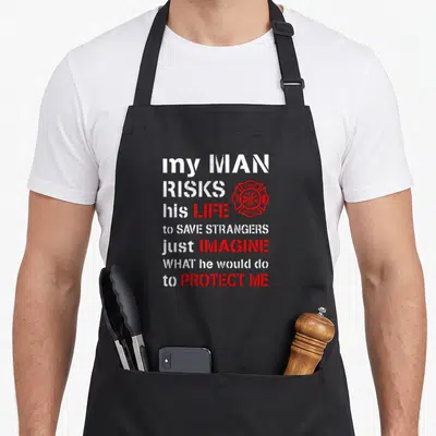 Risk wife apron - Distressed white and red text with red fire dept badge reading my man risks his life — shown on a black cotton canvas apron against a white background