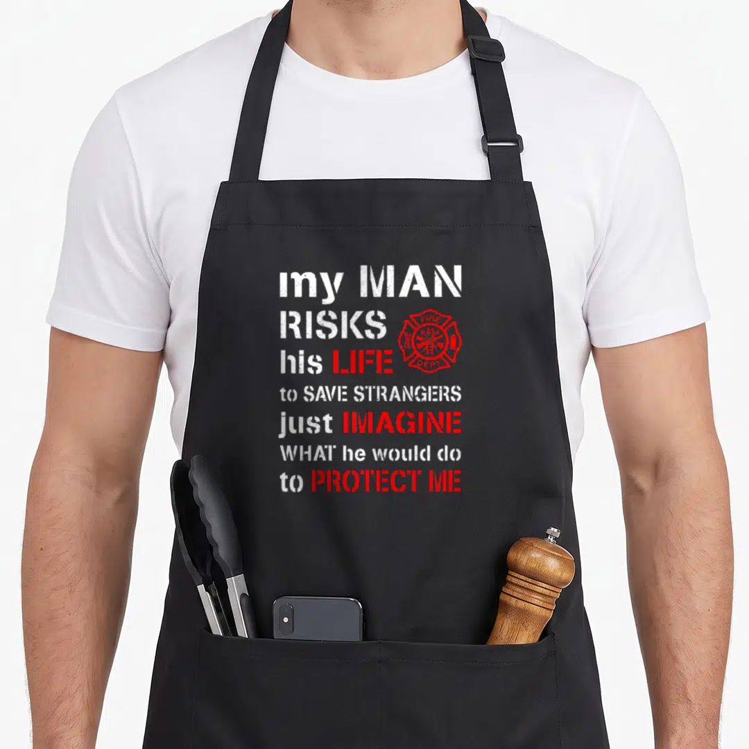 Risk wife apron - Distressed white and red text with red fire dept badge reading my man risks his life — shown on a black cotton canvas apron against a white background