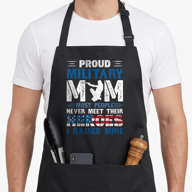 Mine military apron - PROUD MILITARY MOM text with flag HEROES and white female soldier silhouette inside the letter M — shown on a black cotton canvas apron against a white background