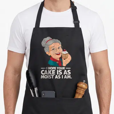 Hope cake apron - Cartoon elderly woman holding cherry cupcake with humorous quote about moisture — shown on a black cotton canvas apron against a white background